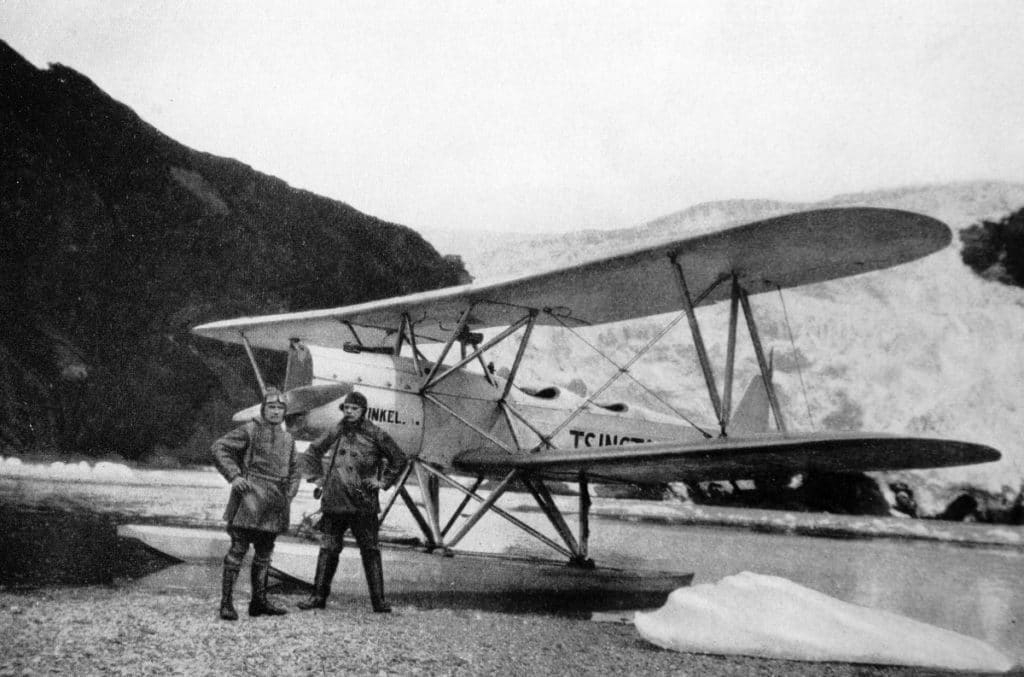 schwarz-weiß Bild von Filmmaterial aus den späten 1920ern: Zwei Männer vor einem Flugzeug